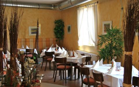 Ein kleiner Restaurantbereich mit mehreren Tischen, die mit weißen Tischdecken und Servietten gedeckt sind, Holzstühlen, Wandkunst und Topfpflanzen an den sonnenbeschienenen Fenstern.