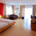 Ein geräumiges Hotelzimmer mit einem Doppelbett, einem Sofa, einem Fernseher, zwei großen Fenstern mit gestreiften Vorhängen, Holzböden und minimalistischer Einrichtung.