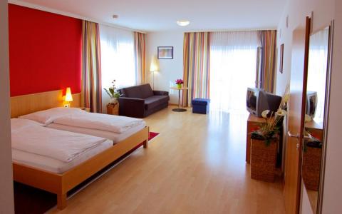 Ein geräumiges Hotelzimmer mit einem Doppelbett, einem Sofa, einem Fernseher, zwei großen Fenstern mit gestreiften Vorhängen, Holzböden und minimalistischer Einrichtung.