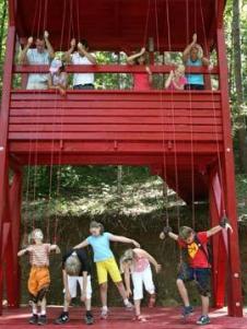 Kinder auf einer unteren Plattform scheinen wie Marionetten von Menschen gesteuert zu werden, die Seile auf einer oberen Plattform einer roten Holzkonstruktion im Freien halten.