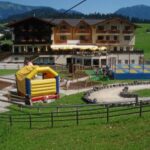 Ein Hotel mit einem großen Spielplatz im Freien mit Hüpfburg, Trampolinen und einer Go-Kart-Bahn vor der Kulisse von Hügeln und Bergen.