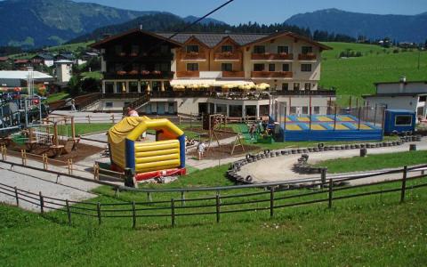 Ein Hotel mit einem großen Spielplatz im Freien mit Hüpfburg, Trampolinen und einer Go-Kart-Bahn vor der Kulisse von Hügeln und Bergen.
