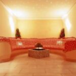 Ein sanft beleuchteter Raum mit einer geschwungenen Mosaikbank, zwei Topfpflanzen und einem zentralen Brunnen auf einem gefliesten Boden.