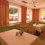 Ein heller Speisesaal mit grün gepolsterten Bänken, Holzstühlen, Tischen mit beigen Tischtüchern und großen Fenstern mit orangefarbenen Vorhängen.