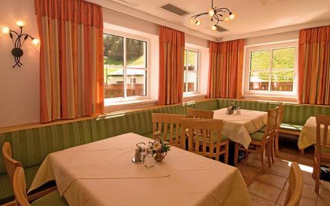 Ein heller Speisesaal mit grün gepolsterten Bänken, Holzstühlen, Tischen mit beigen Tischtüchern und großen Fenstern mit orangefarbenen Vorhängen.