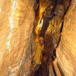 Eine Person in einer gelben Jacke steht in einem engen Durchgang zwischen hohen, strukturierten Höhlenwänden.