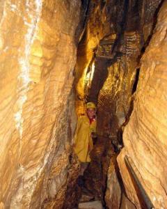 Eine Person in einer gelben Jacke steht in einem engen Durchgang zwischen hohen, strukturierten Höhlenwänden.