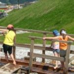 Eine Gruppe von Kindern überquert an einem sonnigen Tag in einem Naherholungsgebiet einen kleinen Holzsteg über einen seichten Bach.