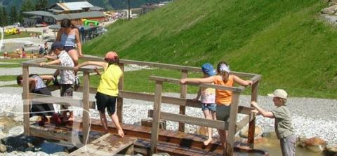 Eine Gruppe von Kindern überquert an einem sonnigen Tag in einem Naherholungsgebiet einen kleinen Holzsteg über einen seichten Bach.