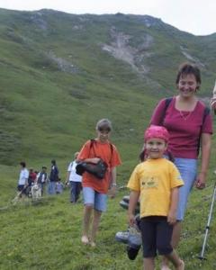 Eine Gruppe von Menschen, darunter Kinder und Erwachsene, geht auf einem grasbewachsenen Hügel mit einem Berg im Hintergrund spazieren.