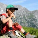Ein Junge mit grüner Mütze und roter Hose sitzt auf einem Felsen und isst eine Kleinigkeit, während im Hintergrund die Berge zu sehen sind.