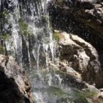 Ein kleiner Wasserfall fließt über moosbewachsene Felsen, die Wassertropfen plätschern und das Sonnenlicht spiegelt sich auf der nassen Steinoberfläche.