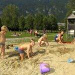 Kinder spielen im Sand auf einem Spielplatz mit Rutsche und Klettergerüst, während sich Erwachsene und andere Kinder im Hintergrund auf einer Wiese entspannen und spielen.