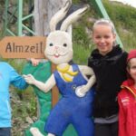 Drei Kinder posieren im Freien neben einer großen bemalten Holzfigur eines Cartoon-Hasen, der eine blaue Latzhose und eine gelbe Fliege trägt, mit einem Schild mit der Aufschrift "Almzeit" im Hintergrund.