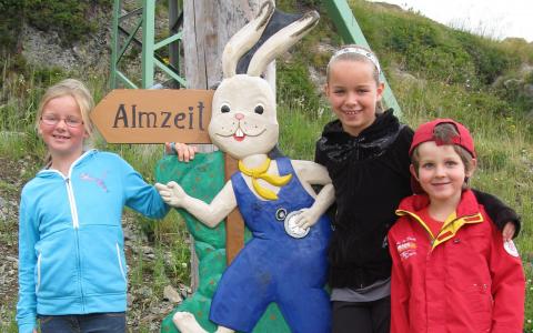 Drei Kinder posieren im Freien neben einer großen bemalten Holzfigur eines Cartoon-Hasen, der eine blaue Latzhose und eine gelbe Fliege trägt, mit einem Schild mit der Aufschrift "Almzeit" im Hintergrund.