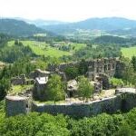 Luftaufnahme einer steinernen Burgruine, umgeben von Bäumen und Feldern, mit Hügeln und Bergen im fernen Hintergrund.