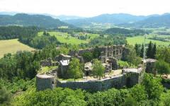 Luftaufnahme einer steinernen Burgruine, umgeben von Bäumen und Feldern, mit Hügeln und Bergen im fernen Hintergrund.