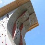 Eine Person klettert an einer künstlichen Kletterwand im Freien mit verschiedenen Griffen, unter einem sonnigen blauen Himmel.