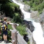 Drei Personen mit Rucksäcken stehen auf einem Pfad hinter einem Holzzaun und blicken auf einen schnell fließenden Fluss, der von Felsen und Grün umgeben ist.