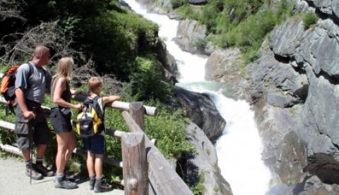 Drei Personen mit Rucksäcken stehen auf einem Pfad hinter einem Holzzaun und blicken auf einen schnell fließenden Fluss, der von Felsen und Grün umgeben ist.