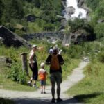 Eine Familie mit zwei Kindern geht auf einem Weg durch grüne Hügel in Richtung einer Steinbrücke und eines Wasserfalls im Hintergrund.