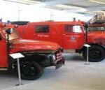 In einem Museum sind mehrere alte rote Feuerwehrautos ausgestellt, vor denen jeweils Schilder mit einer Beschreibung der Fahrzeuge angebracht sind.