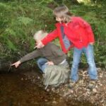Zwei Kinder an einem Bach, eines hockt und beide zeigen auf etwas im Wasser, umgeben von Grün und Steinen.
