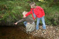 Zwei Kinder an einem Bach, eines hockt und beide zeigen auf etwas im Wasser, umgeben von Grün und Steinen.