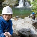 Ein Kind mit weißem Schutzhelm und blauem Kapuzenpulli hält Steine an einem Bach, während ein anderes Kind mit einem blauen Helm im Hintergrund in der Nähe eines Wasserfalls zu sehen ist.