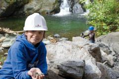 Ein Kind mit weißem Schutzhelm und blauem Kapuzenpulli hält Steine an einem Bach, während ein anderes Kind mit einem blauen Helm im Hintergrund in der Nähe eines Wasserfalls zu sehen ist.