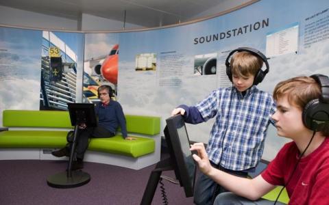 Zwei Jungen benutzen Touchscreen-Monitore mit Kopfhörern an einem interaktiven Exponat namens "Soundstation", während ein Mann im Hintergrund sitzt und ebenfalls Kopfhörer trägt.