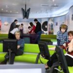 Menschen verschiedenen Alters interagieren mit Computern und Exponaten in einem modernen, geschwungenen Raum mit grünen Sitzgelegenheiten und Informationsdisplays an den Wänden.