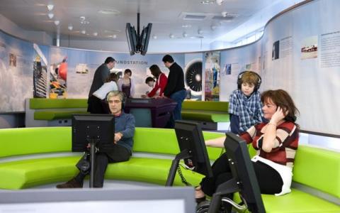 Menschen verschiedenen Alters interagieren mit Computern und Exponaten in einem modernen, geschwungenen Raum mit grünen Sitzgelegenheiten und Informationsdisplays an den Wänden.