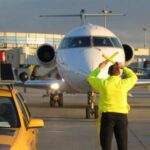 Ein Mitarbeiter des Bodenpersonals eines Flughafens lenkt ein rollendes Flugzeug mit Hilfe von Rangierstangen, während ein anderer Mitarbeiter und ein gelbes Flughafenfahrzeug in der Nähe sind.