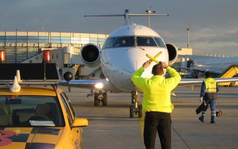 Ein Mitarbeiter des Bodenpersonals eines Flughafens lenkt ein rollendes Flugzeug mit Hilfe von Rangierstangen, während ein anderer Mitarbeiter und ein gelbes Flughafenfahrzeug in der Nähe sind.