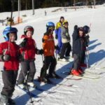 Eine Gruppe von Kindern und ein Erwachsener stehen in einer Reihe auf einer verschneiten Skipiste, tragen Helme und Skiausrüstung und halten Skistöcke.