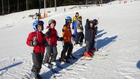 Eine Gruppe von Kindern und ein Erwachsener stehen in einer Reihe auf einer verschneiten Skipiste, tragen Helme und Skiausrüstung und halten Skistöcke.