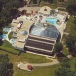 Luftaufnahme einer Freizeitanlage mit Außen- und Innenpools, Whirlpools, einer Glaskuppel, Bäumen und offenen Grasflächen.