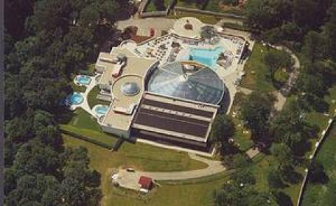 Luftaufnahme einer Freizeitanlage mit Außen- und Innenpools, Whirlpools, einer Glaskuppel, Bäumen und offenen Grasflächen.