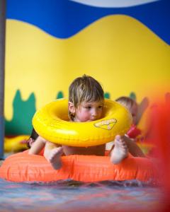 Ein Kind in einem Pool trägt einen gelben Schwimmring um den Hals und liegt auf einem orangefarbenen Schwimmkörper. Im Hintergrund sind bunte, abstrakte Formen zu sehen.