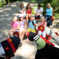 Im Vordergrund sind zwei Sockenpuppen zu sehen, während im Hintergrund eine Gruppe von sieben Personen im Freien sitzt und unscharf ist.