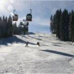 Zwei Personen fahren an einem sonnigen Tag eine verschneite Piste mit Kiefern an den Seiten und Skiliften über sich hinunter.