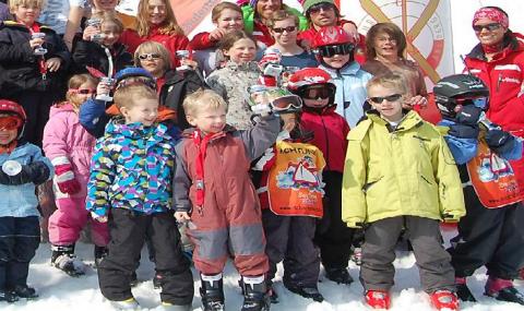 Eine Gruppe von Kindern und Erwachsenen in Winter- und Skikleidung posiert gemeinsam im Schnee, einige mit Getränken in der Hand, vor einer Kulisse mit Skimotiven.