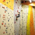 Eine Person klettert an einer Indoor-Felswand mit bunten Griffen, trägt Sicherheitsausrüstung und einen Klettergurt. Oben ist ein digitaler Timer zu sehen.