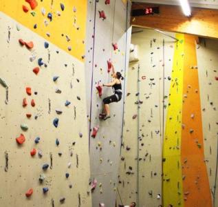 Eine Person klettert an einer Indoor-Felswand mit bunten Griffen, trägt Sicherheitsausrüstung und einen Klettergurt. Oben ist ein digitaler Timer zu sehen.