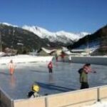 Menschen laufen Schlittschuh auf einer schneebedeckten Eisbahn im Freien, im Hintergrund sind Berge und Häuser unter einem klaren blauen Himmel zu sehen.