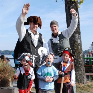 Zwei Erwachsene und drei Kinder in Piratenkostümen posieren und lächeln im Freien in der Nähe eines Baumes und eines Gewässers.