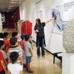 Eine Frau zeigt auf eine Karte an der Wand, während eine Gruppe von Kindern in einem Museum vor ihr steht.