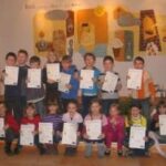 Eine Gruppe von Kindern posiert in einem Raum, jedes hält eine Urkunde in der Hand, während hinter ihnen bunte Kunstwerke an der Wand hängen.