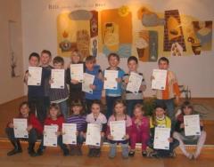 Eine Gruppe von Kindern posiert in einem Raum, jedes hält eine Urkunde in der Hand, während hinter ihnen bunte Kunstwerke an der Wand hängen.